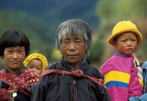 In Bhutan, one older woman and one younger woman with young child strapped to back, another young child held on hip by person of