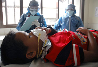 Young boy lies in hospital bed, holds protective mask to face, nurses observe in background