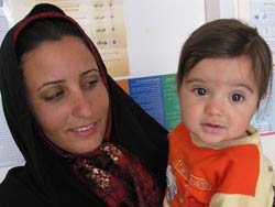 A woman with a headscarf on holds and looks at a baby, the baby looks at the camera, both are in front of a bulletin board