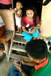 Man seated takes notes on handheld device, woman sits on steps in doorway, toddler stands behind her, drinks from bottle