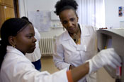 Female medical instructor looks on as female medical student works on computer equipment, both in lab coats