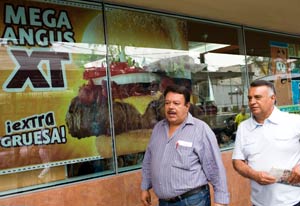 Photo: Large sign picturing fast food hamburger reading Mega Angus XT in window on street, two men walk past