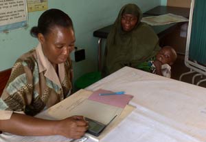 Woman seated at desk enters data into handheld device, seated next to desk woman holds a young child in her arms