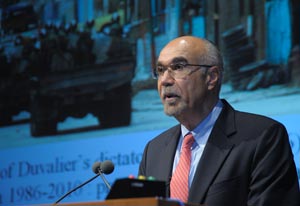 Photo: Dr. Pape speaks into a microphone at a podium, a slide projected in backgound