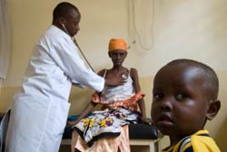 Photo: Young boy, seated in foreground, looks at camera, doctor in lab coat listens to emaciated woman's chest with stethoscope