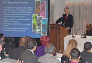 Dr Francis Collins speaks at a podium in front of full conference room, slide projected to his right