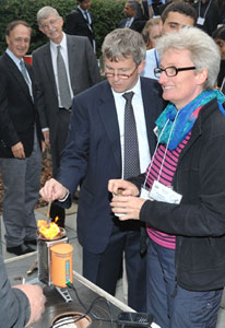 Man and woman test small, lit cookstove in foreground, Dr Francis Collins looks on in background