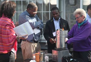 Woman adjusts lit metal cookstove, three people look on to observe