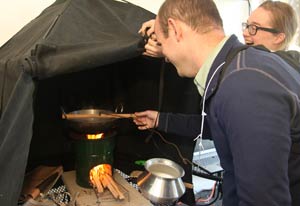 Man holds instrument to measure emissions over lit wood-burning stove, which burns under a hood, woman holds up hood