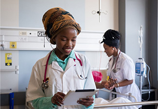 Two female doctors in the hospital, one looking at a tablet and the other tending to a patient.