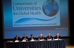 Row of 6 men seated at a panel table at the front of a conference room under a projected slide reading Consortium of Universities for Global Health